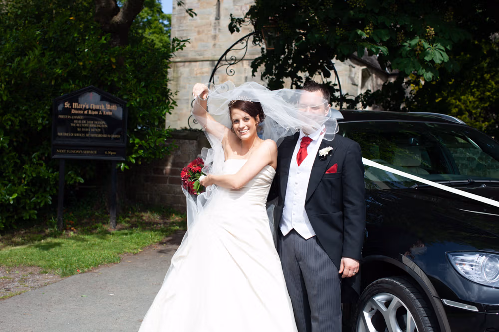 Bride and groom laughing as the wind lifts her veil beside their wedding car at St Mary’s Church in Wath.