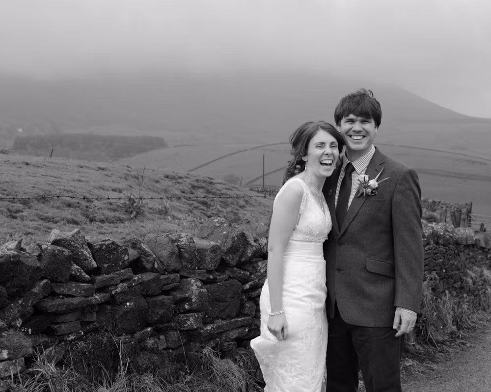 Wedding couple laughing together on a narrow road on Pendle Hill, walking through light rain under dramatic skies in monochrome.