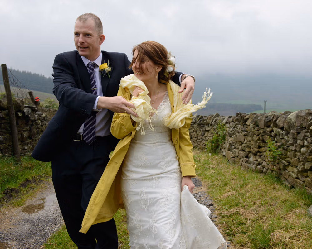 A groomsman gently helps the bride adjust her raincoat and scarf as they walk back towards the car on a wet country road, a quiet moment of care after portraits on Pendle Hill.