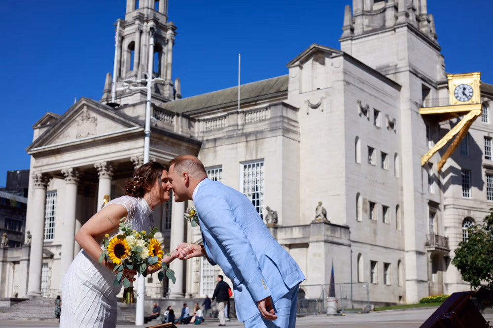 Bride and groom leaning nose to nose over a piano near Leeds Civic Hall, smiling in bright sunshine.