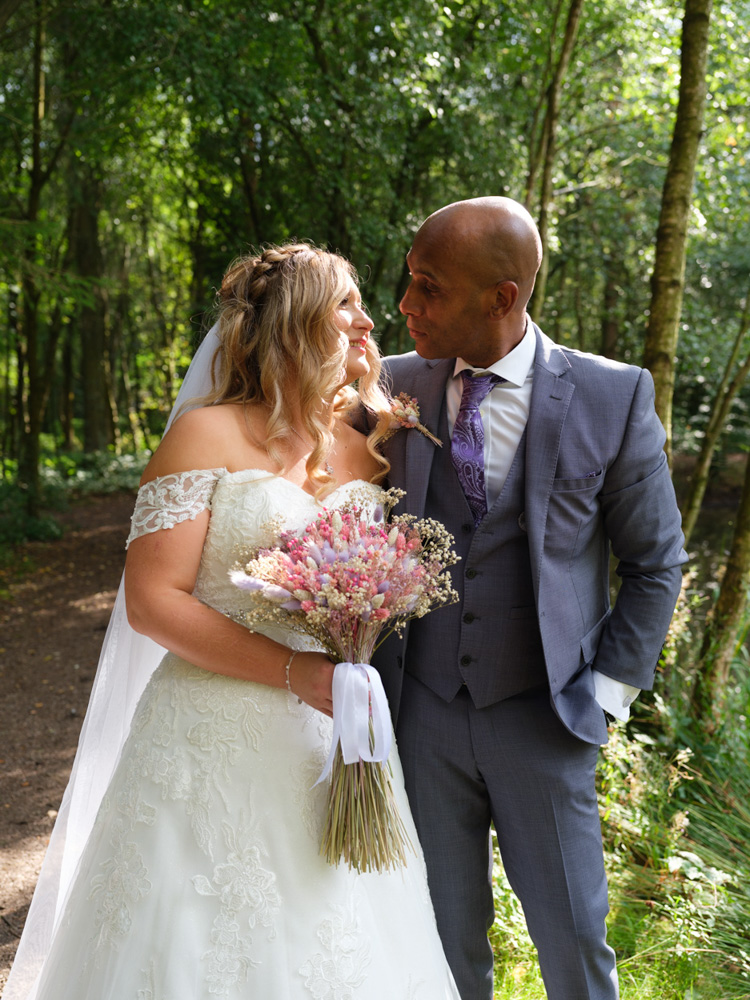 Feydrah and Dean sharing a quiet glance beneath dappled woodland light at Chevin Country Park Hotel during their summer wedding.