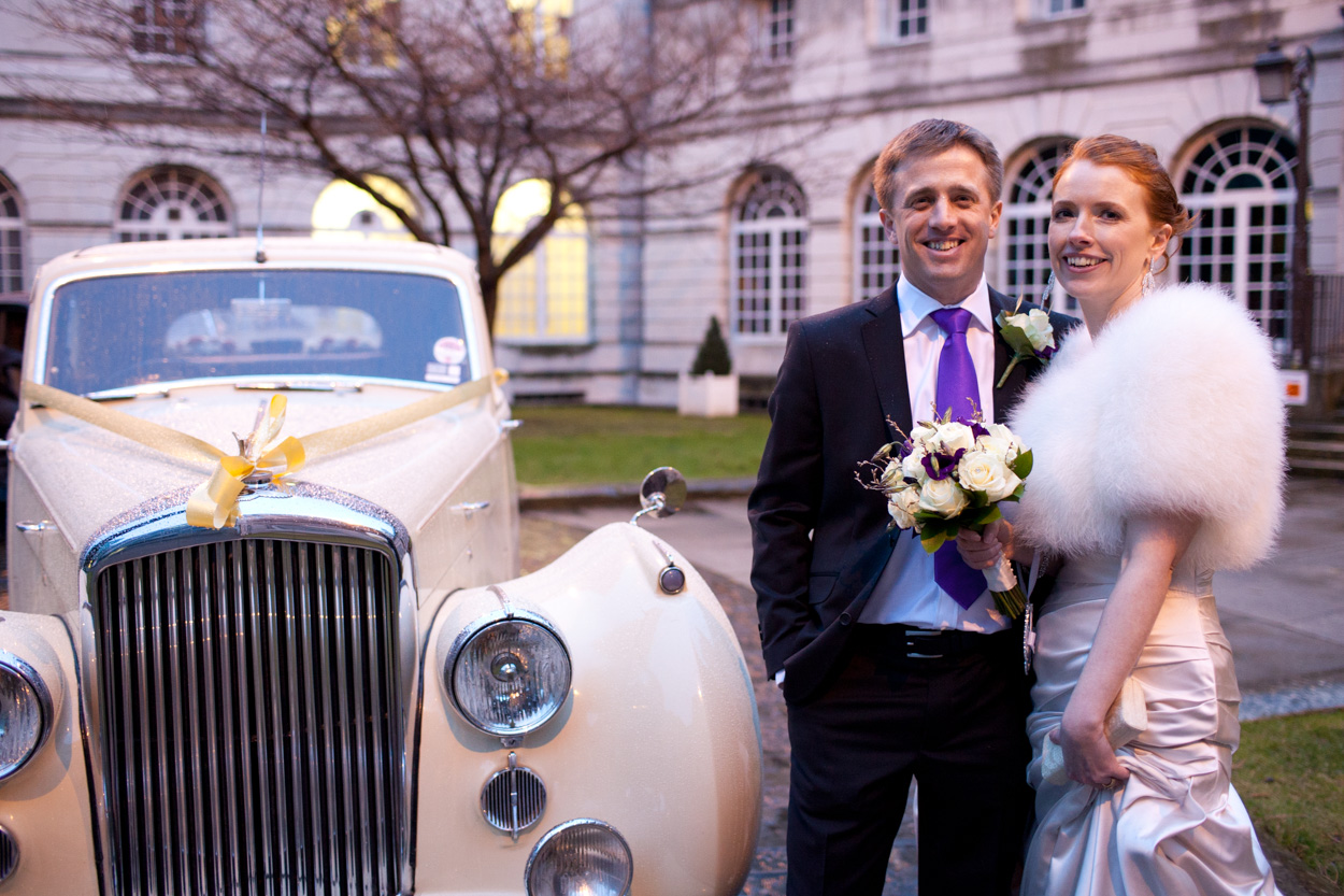 Sarah & Mark smile beside their cream vintage wedding car outside Leeds Civic Hall as light February rain falls before heading to The Headingley Experience.