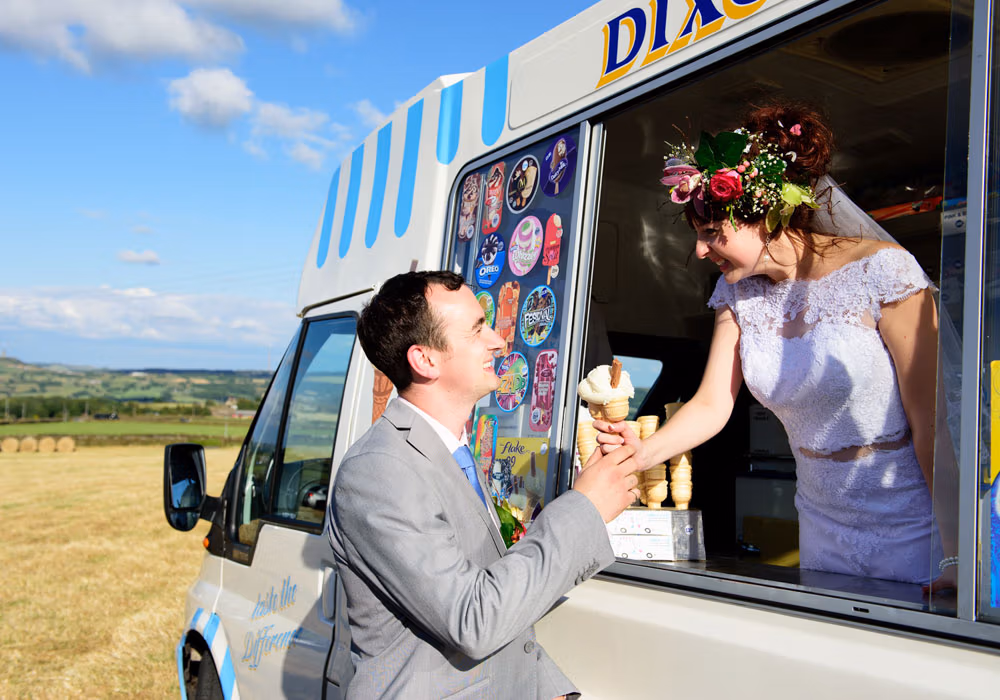 Bride Chloe inside their wedding ice-cream van handing John a 99-Flake ice cream with beautiful sunshines and blue skies behind.Their DIY home field wedding reception.