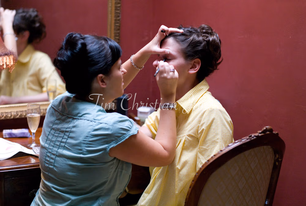 The bride’s sister leans in to apply makeup with care and closeness.