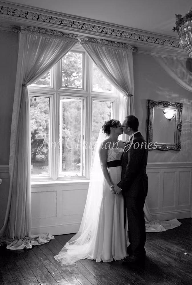 Julie and Pat share a kiss by the tall window of Woodborough Hall, framed in rich fabric and polished floors.