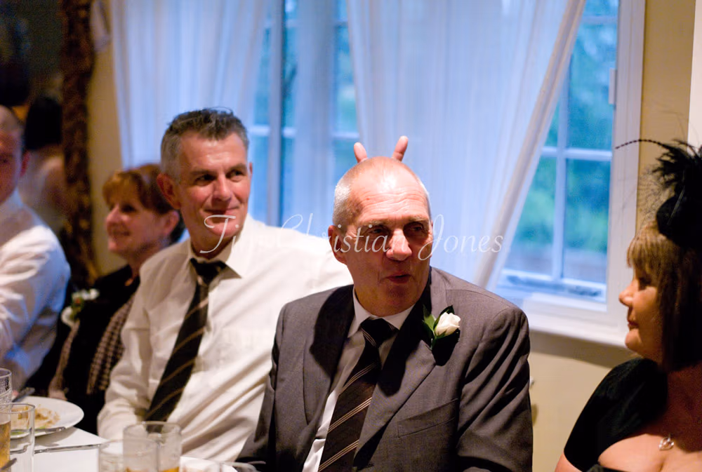 Candid moment at the top table as Julie’s father makes playful rabbit ears behind a guest.