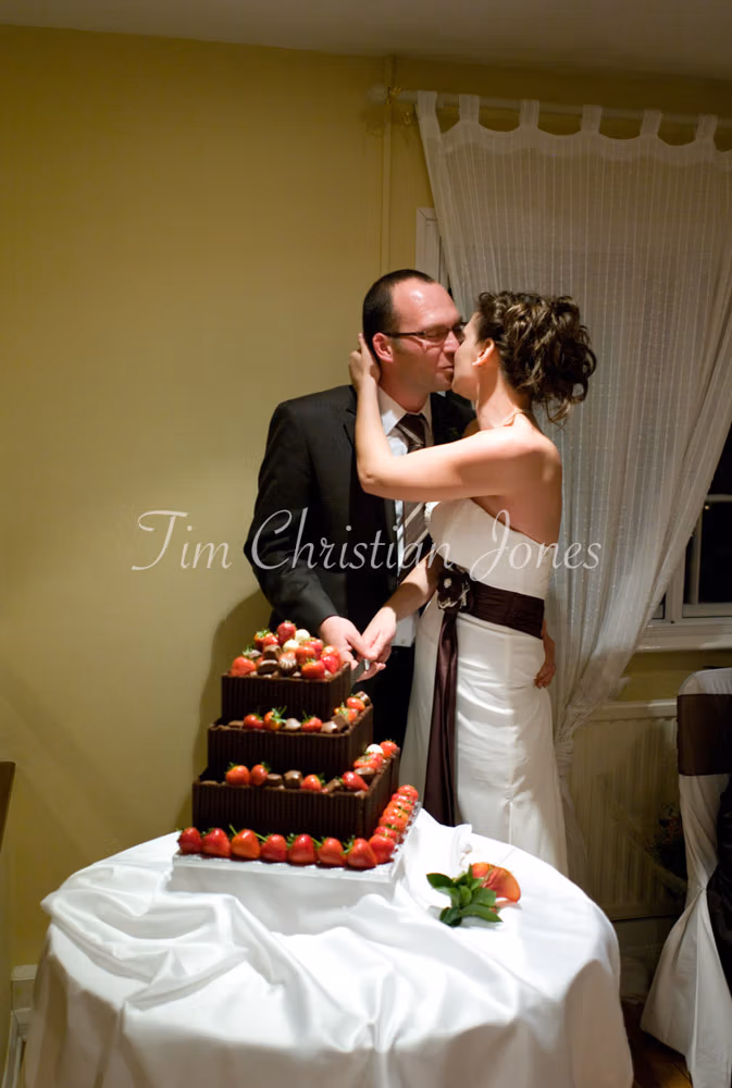 Julie and Pat kiss over the cake knife, hands together on the handle over soft white cloth.