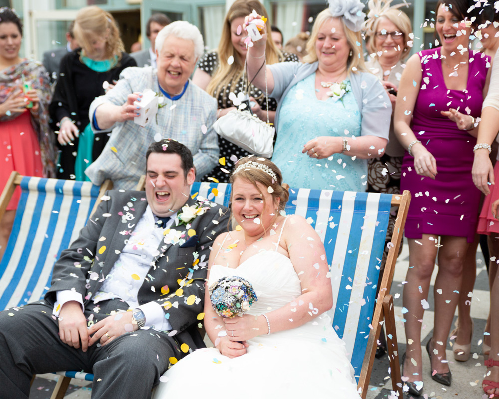 Lisa & Oliver share a joyful moment on the striped deckchairs at Scarborough Spa as family and friends celebrate with confetti behind them. A relaxed seaside wedding moment captured naturally.