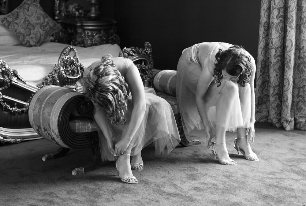 An Allerton Castle wedding near Harrogate — bridesmaids fastening their shoes while seated on the chaise at the foot of the four-poster bed in the bridal suite, captured in timeless monochrome.