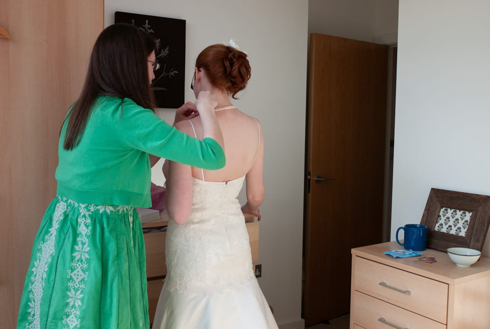 Bride’s best friend fastening faux pearls in apartment, bridesmaid in green, bride with red hair smiling.