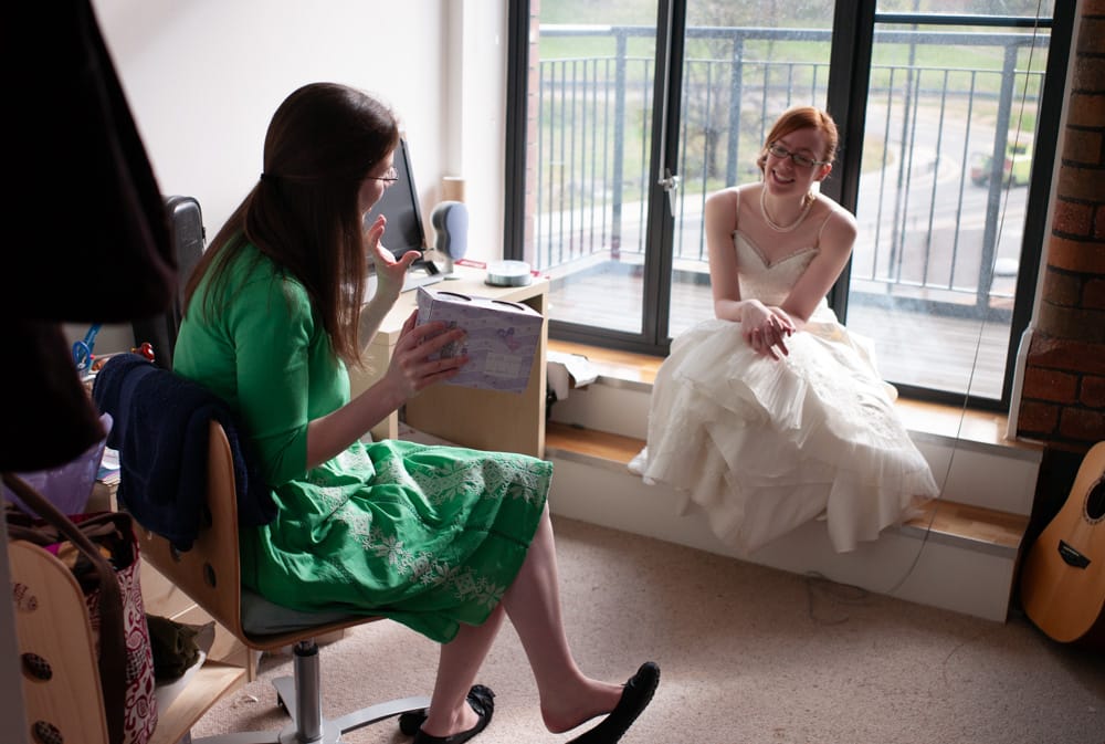 Bride with red hair smiling on window steps, chatting with bridesmaid before wedding.