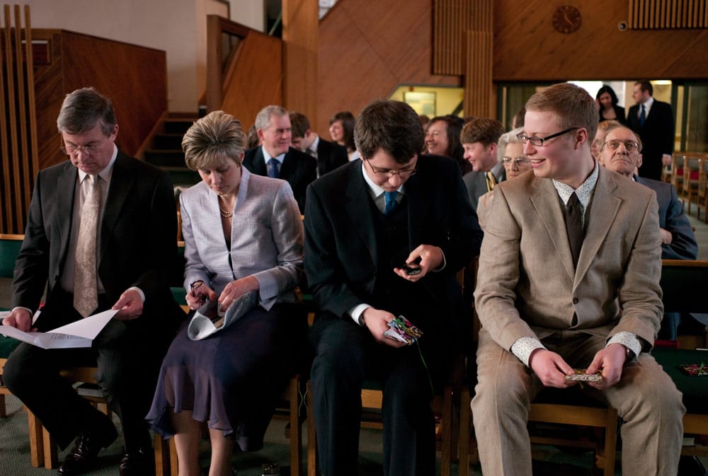 Groom and best man seated at St George’s, checking phone, parents nearby smiling.