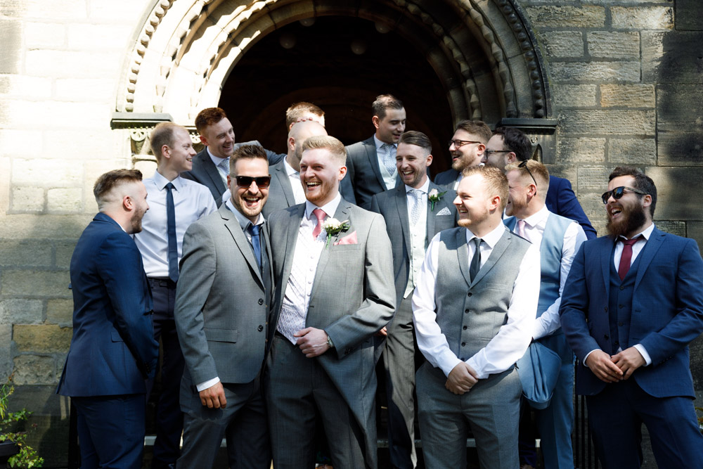 Groom and groomsmen laugh together outside St Oswald’s Church, relaxed and ready for the day ahead.