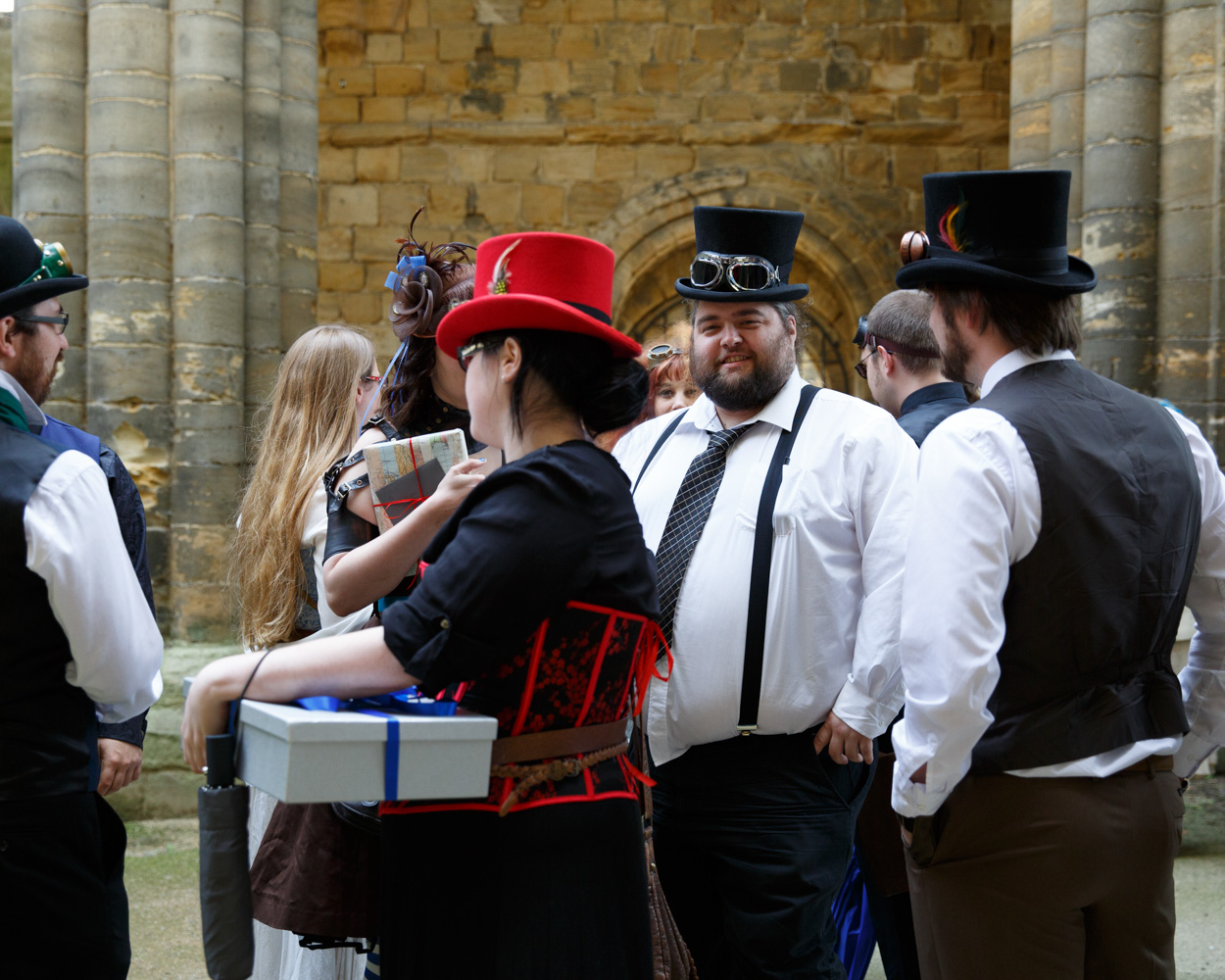 Guests in black, blue, and red top hats chat together before the ceremony at Kirkstall Abbey