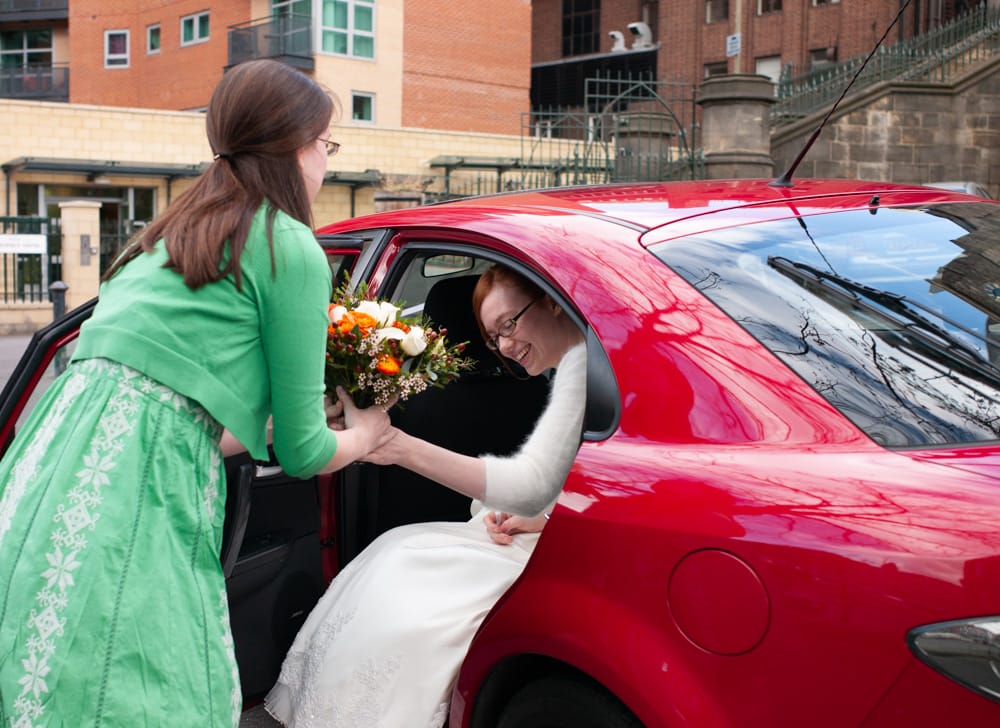 Bride in red car, passing colourful bouquet of white, green and orange to friend.