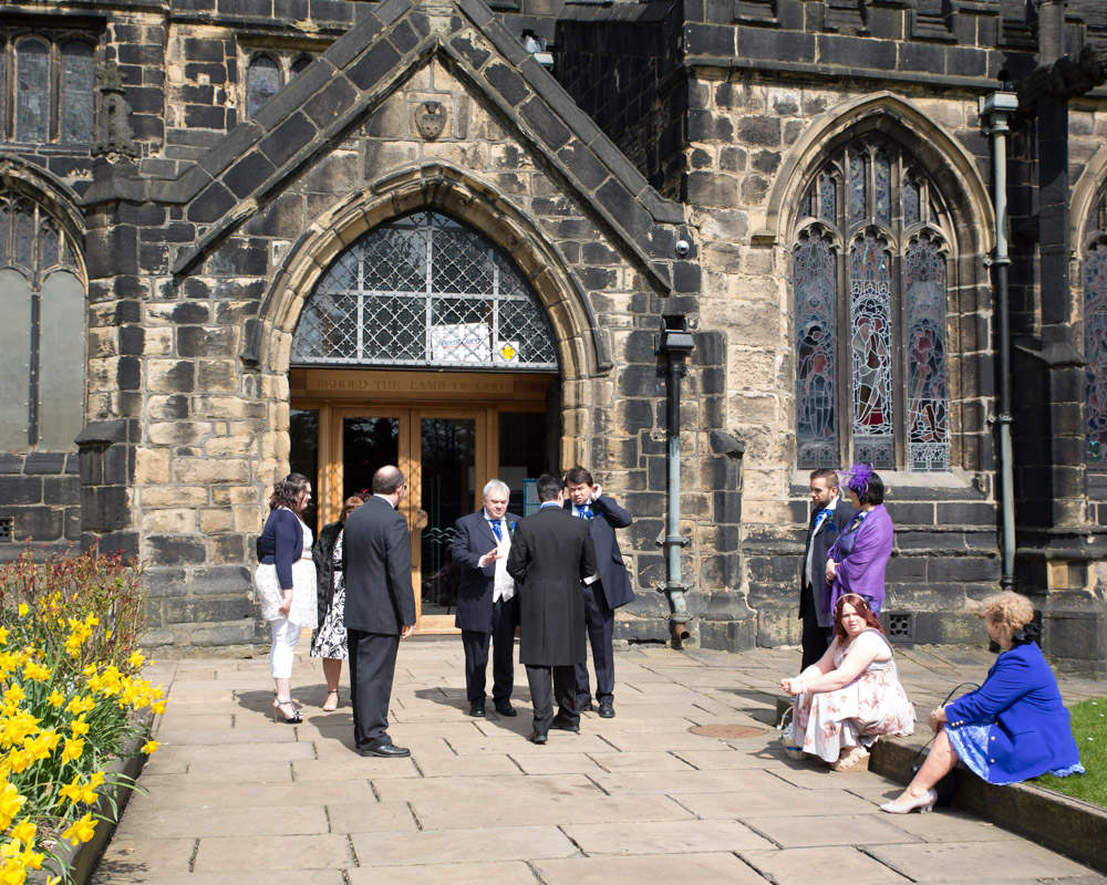A small group of guests gather outside the minster entrance under April sun and daffodils.