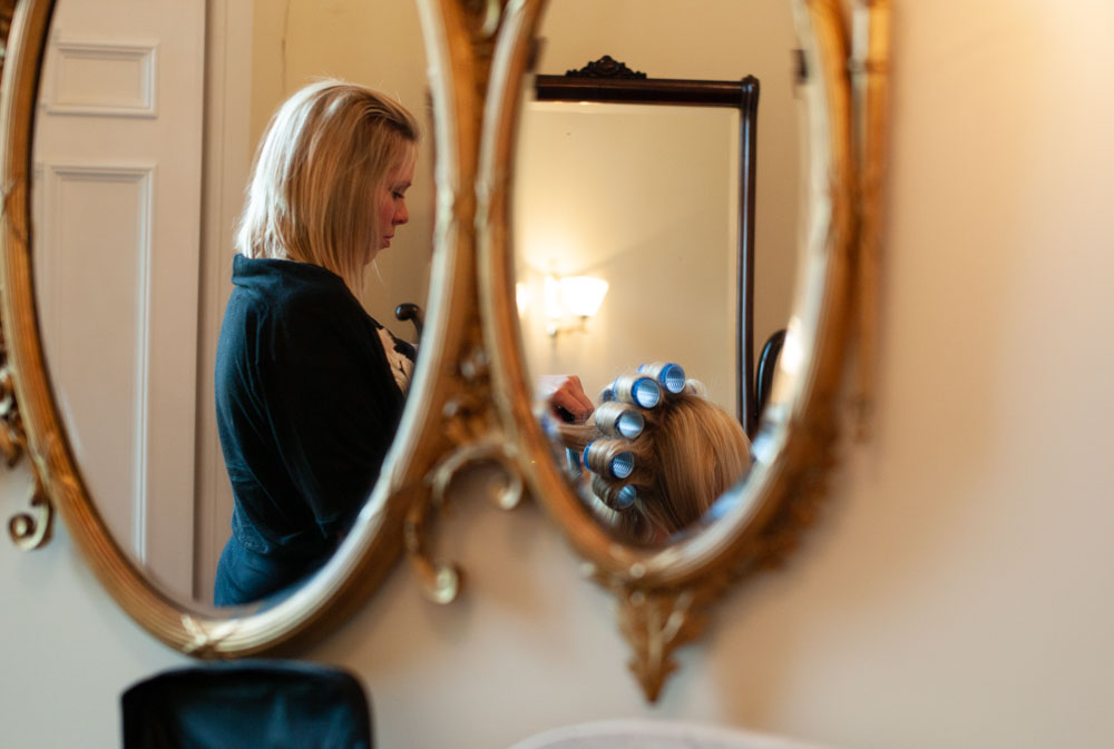 Bride in curlers reflected in twin gold-trimmed oval mirrors as the hairdresser works