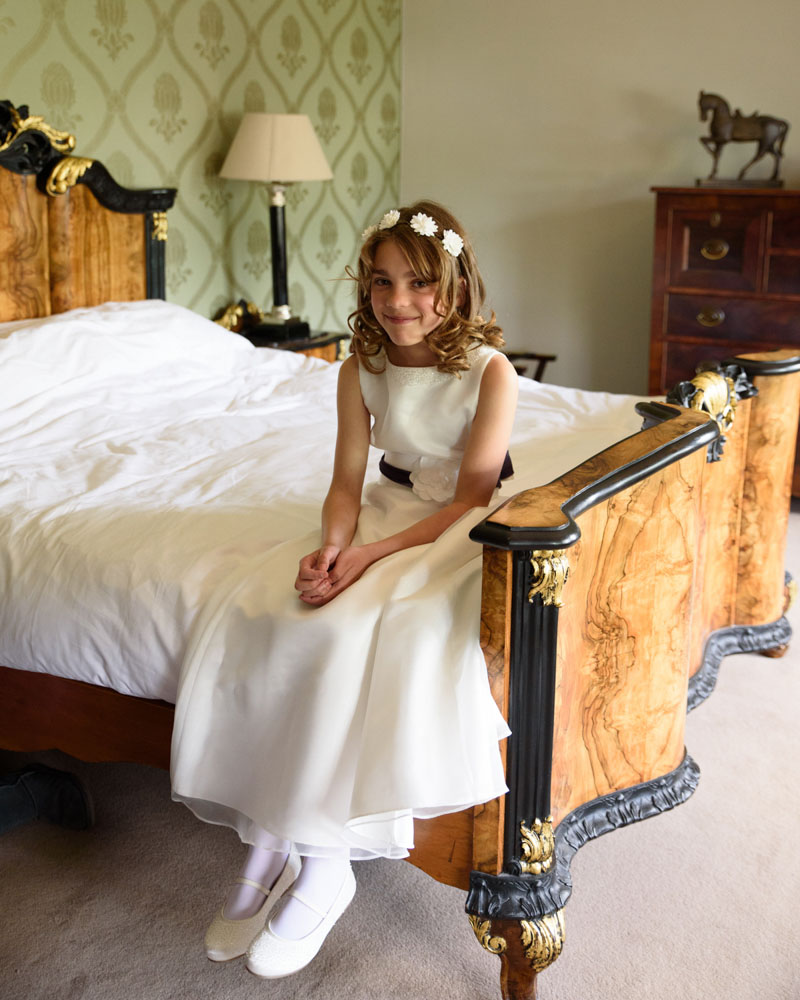 Young bridesmaid sits on the bed in the bridal suite, smiling at the camera.