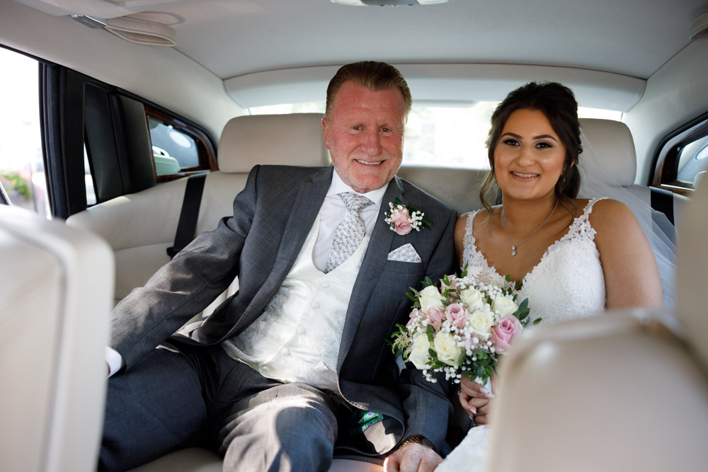 Bride and father in the back of the Rolls-Royce outside St Oswald’s Church, bouquet held gently, both smiling.