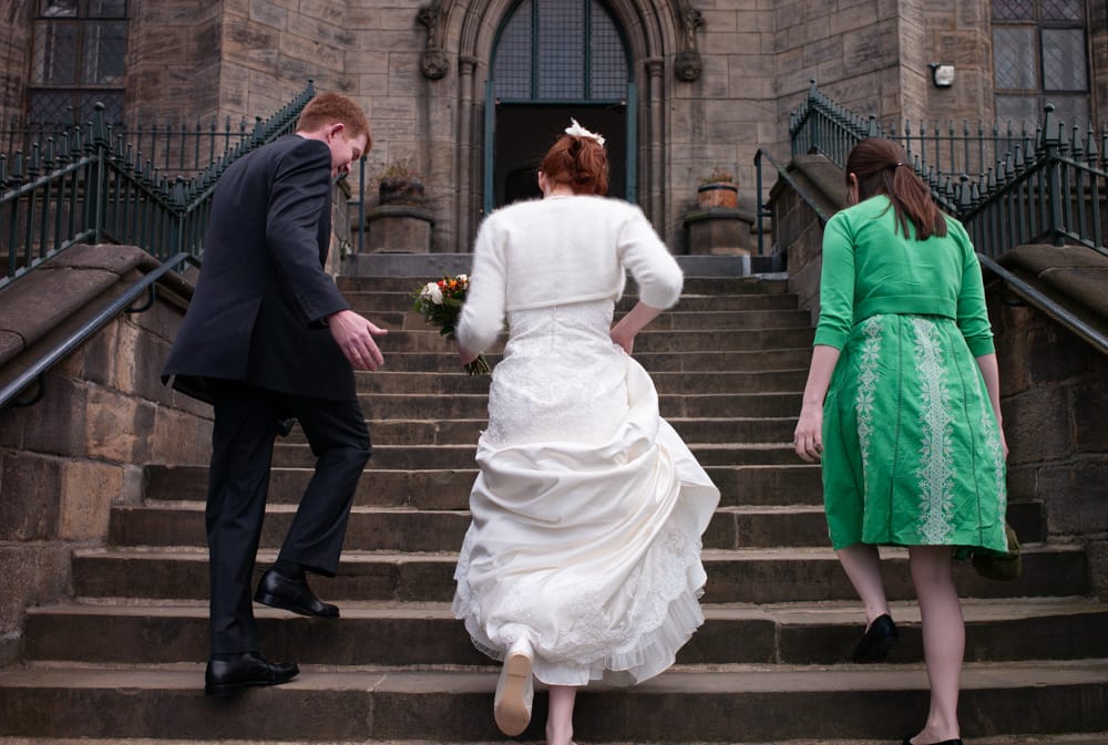 Bride with red hair lifting dress, father and bridesmaid walk up steps into St George’s Church.