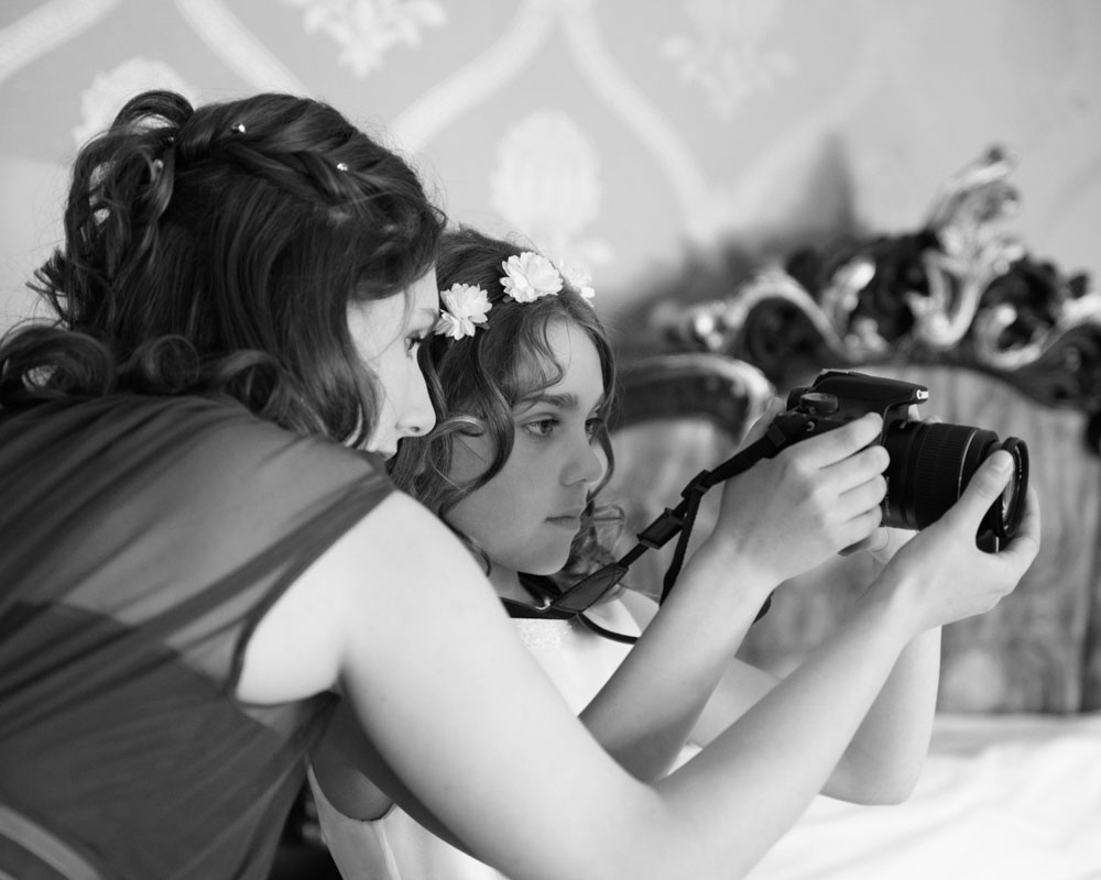Bridesmaid helps her sister with a camera during wedding morning prep.
