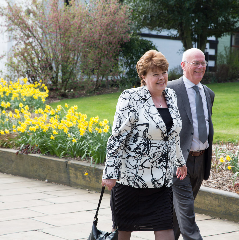 Two smiling guests walk toward Halifax Minster, daffodils blooming behind them.