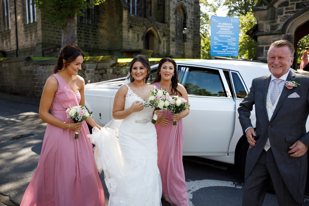 Bride exits the car at St Oswald’s Church, assisted by bridesmaids and her father, the historic church behind them.