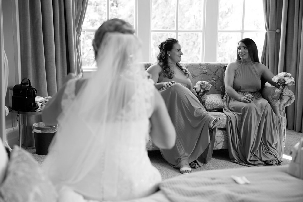 Bride seated on the bed in her veil, looking towards her bridesmaids chatting on the sofa ahead