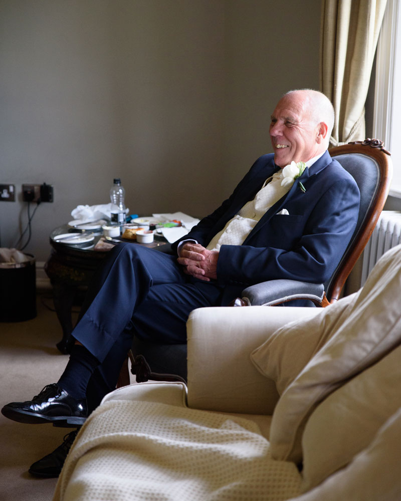 Bride’s father sits smiling in a comfy chair in the bridal