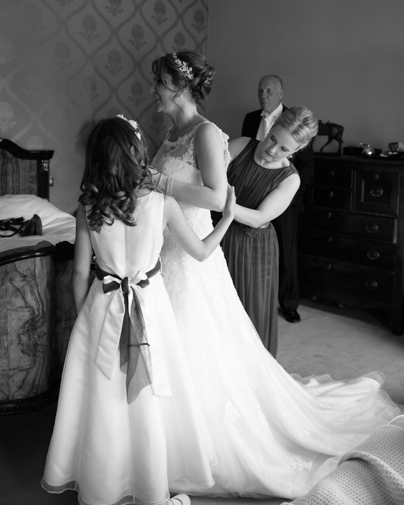 Bride’s sister fastens the dress as the young bridesmaid lends a hand.