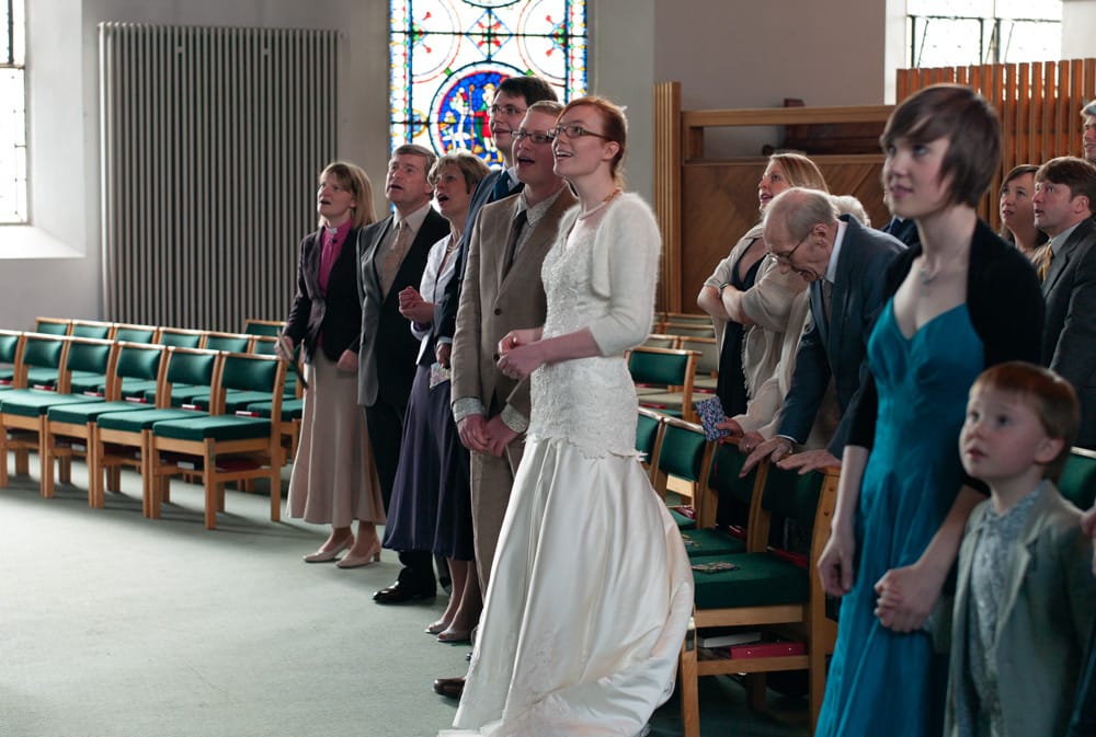 Bride, groom and guests singing together during church ceremony, looking up at words.