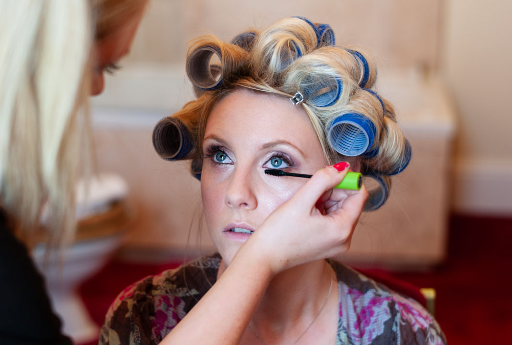 Front view of bride with eyes open as makeup artist steadies her hand.