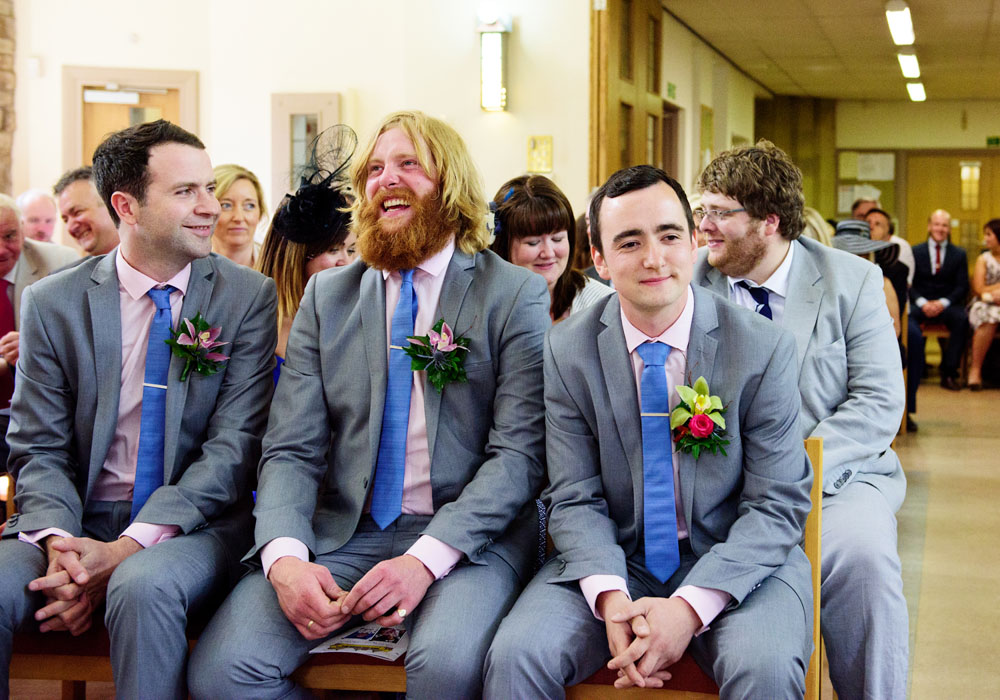Groom and best men smiling, seated at the front row inside St Paul’s Church in Huddersfield.
