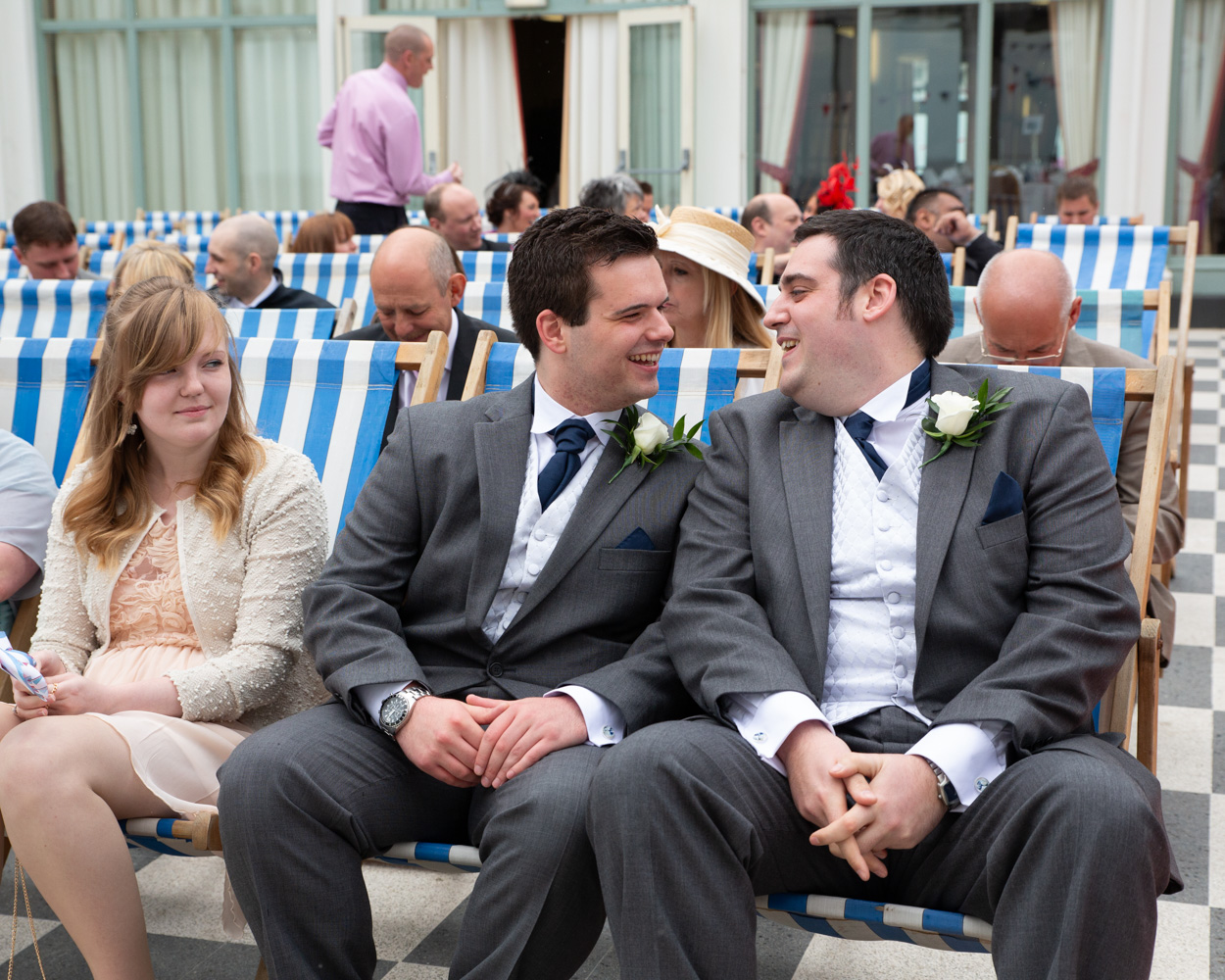 Candid moment of the groom and best man smiling at each other, seated among guests before the ceremony.
