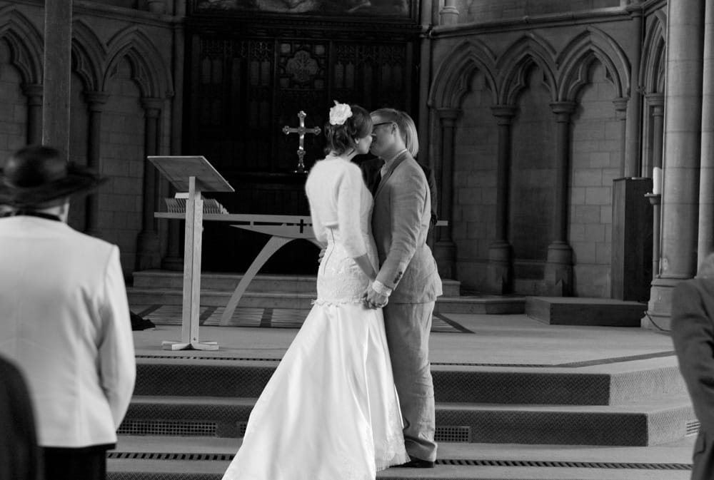 Bride and groom’s first kiss at altar, black and white, holding hands.