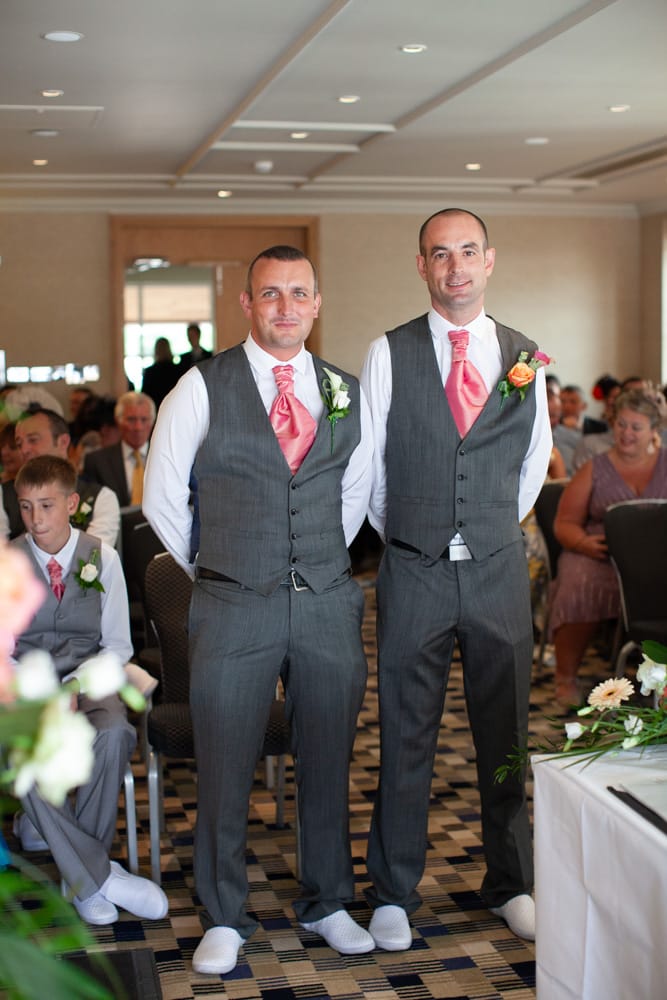 Groom Dave and his best man smile, wearing white shoes and pink cravats, while guests wait for the bride.