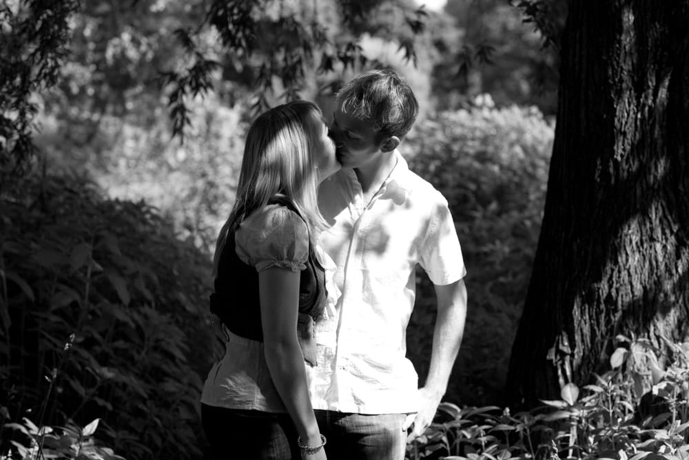Couple kissing in the dappled woodland light among the trees at Kirkstall Abbey, captured in black and white.