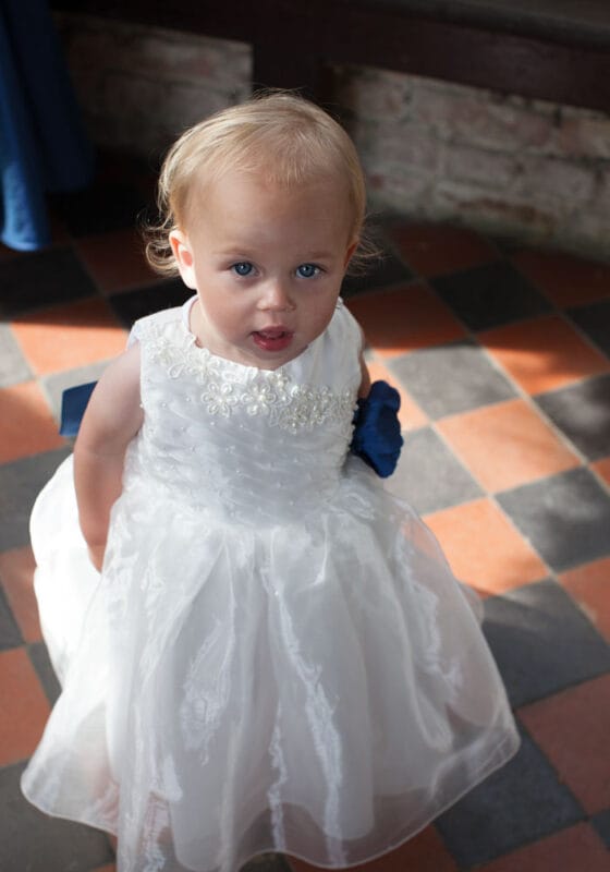 Little bridesmaid looking up with wide eyes on a chequered tiled floor, bathed in soft natural light