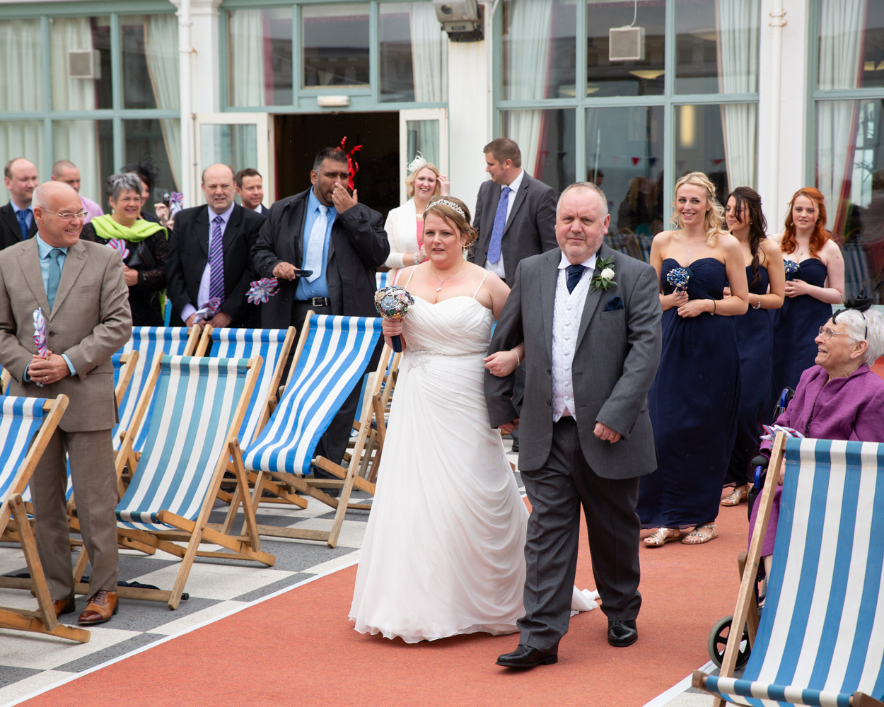 Bride entering the ceremony at Scarborough Spa with her father, bridesmaids following behind smiling.