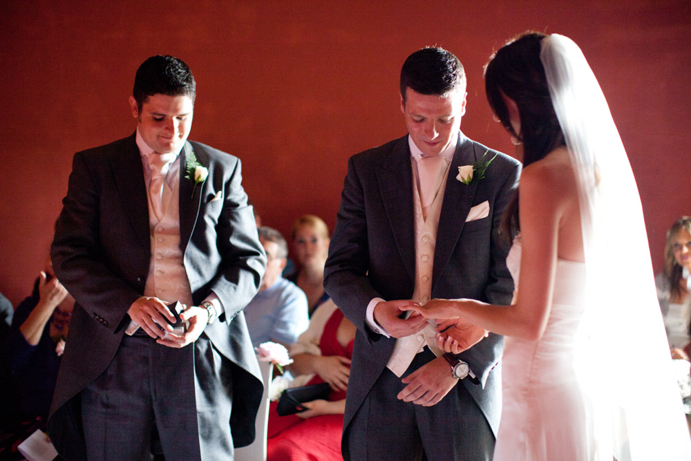 Groom places the wedding ring on the bride’s finger as best man holds the ring box