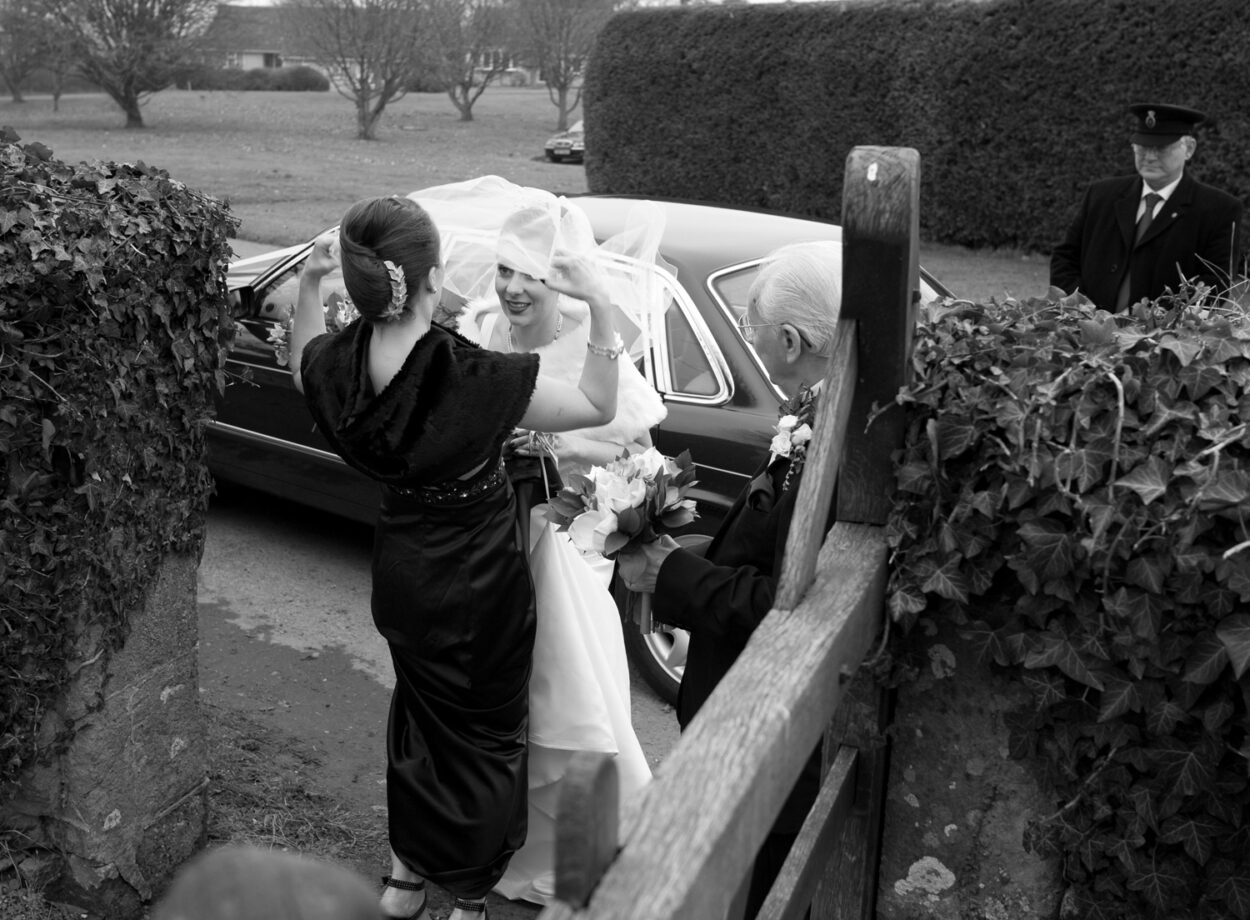Emelie’s sister adjusts her veil at the church gate as grandad holds the bouquet.