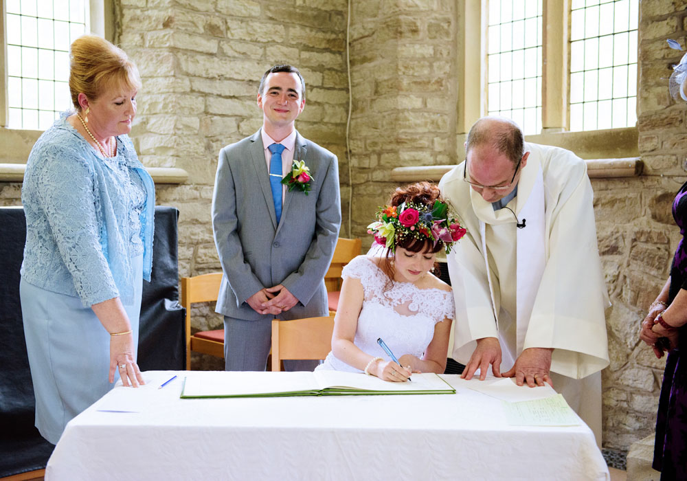 Bride signs the register at St Paul’s Church as the groom smiles warmly at the camera.