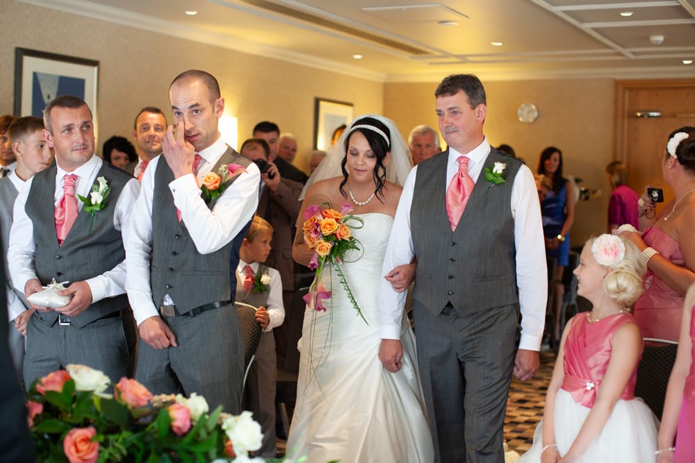 Gemma reaches Dave, holding her orange bouquet, while a little bridesmaid smiles in pink and white.