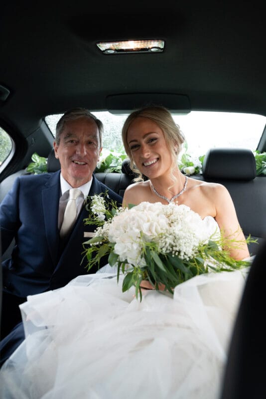 Bride and her father share a smile in the wedding car, bouquet of white flowers and ferns.