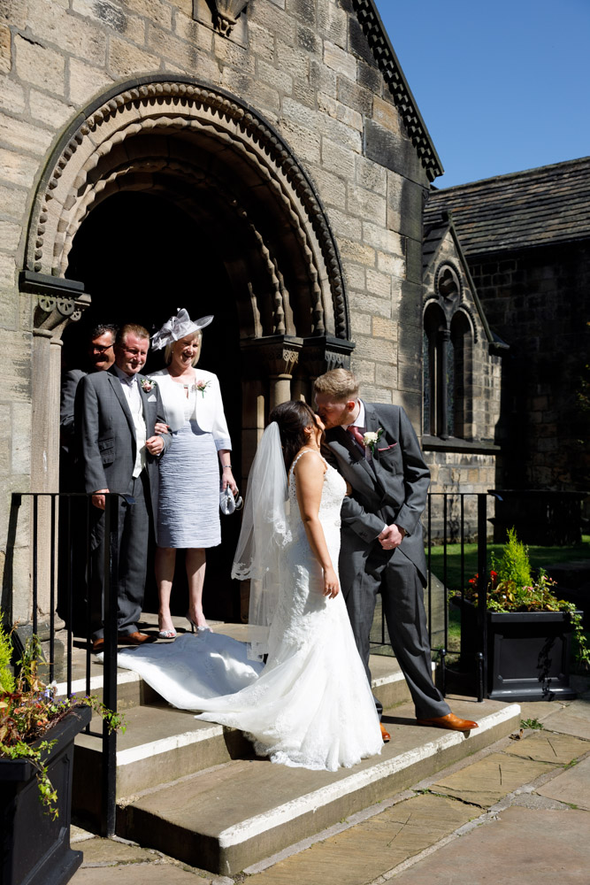 Kyra and Matt pause for a kiss on the stone steps of St Oswald’s Church in Guiseley, with smiling parents close behind.
