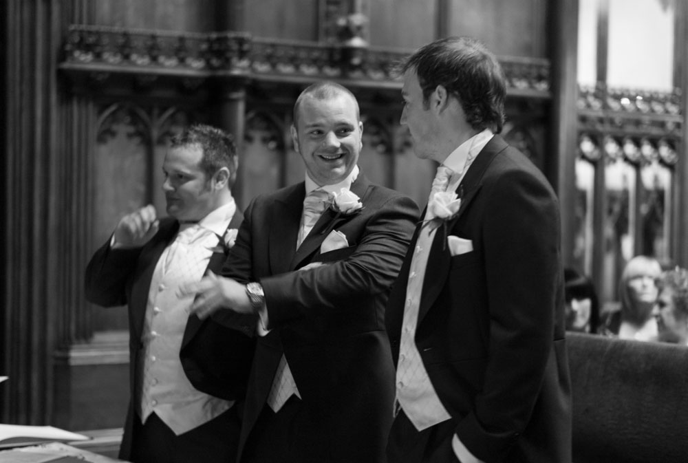 In black and white, the groom and best men smile as they wait at the front of the ceremony room.