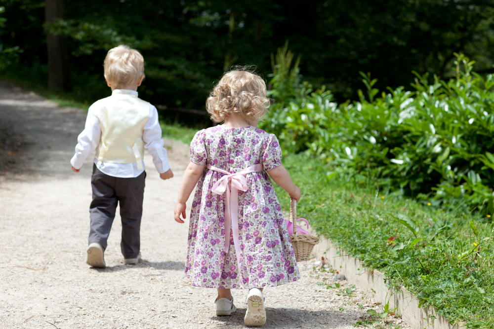 Flower girl walks with the couple’s son along a garden path, basket in hand