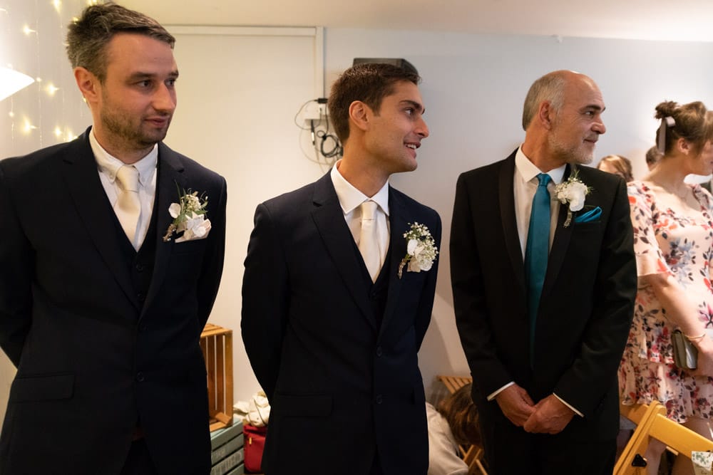 Groom, best man, and father of the groom glance back as bride enters the Chilli Barn with her father.