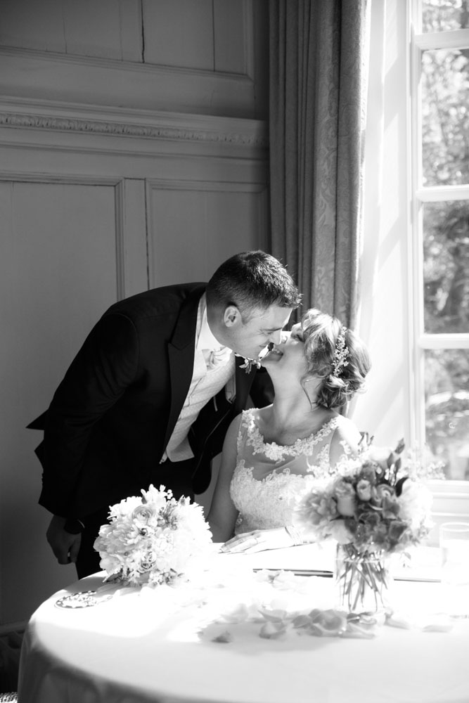 The couple smiles warmly, about to kiss near the bay window.