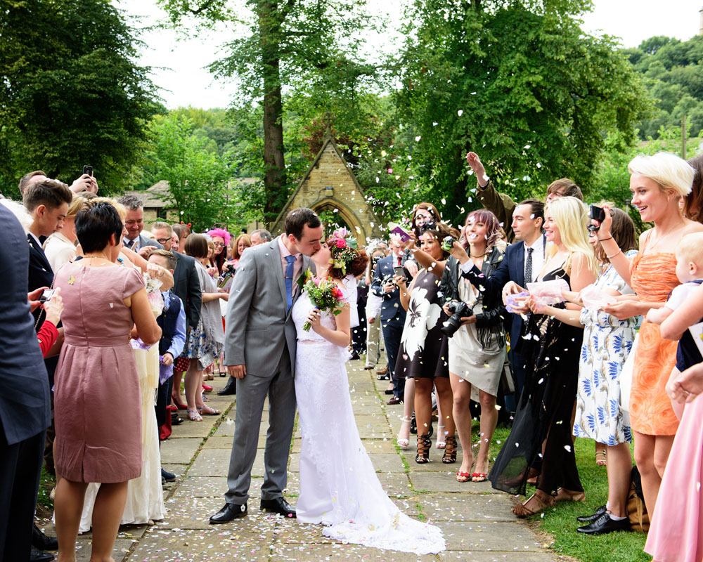 Couple kiss in front of St Paul’s Church gates as confetti flies — framed by trees and guests.